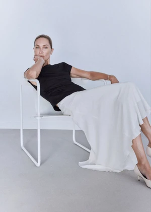 Model Erin Wasson seated in a modern white chair wearing a black cap-sleeve top and flowing white maxi skirt.