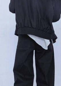 Back view of a model wearing a black bomber jacket and matching black trousers with a white top hem against a seamless backdrop.