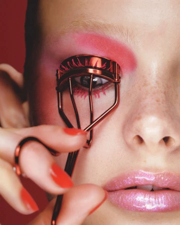 Close-up portrait of a person using a red eyelash curler, featuring bold pink eyeshadow and glossy lips against a red backdrop.
