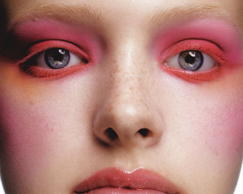 Extreme close-up of a woman's face featuring vibrant pink and coral eyeshadow, glossy pink lips, and blue eyes.