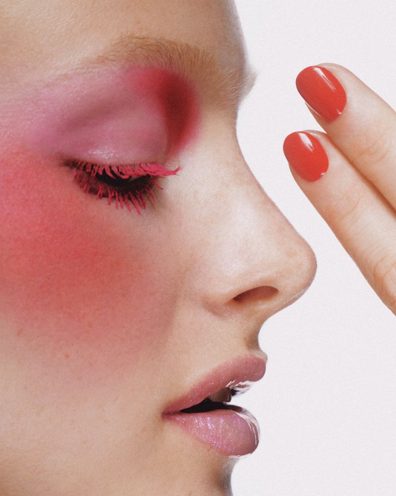 Close-up beauty shot of fair-skinned adult female with striking pink eye makeup and red nails against a clean, bright white background.