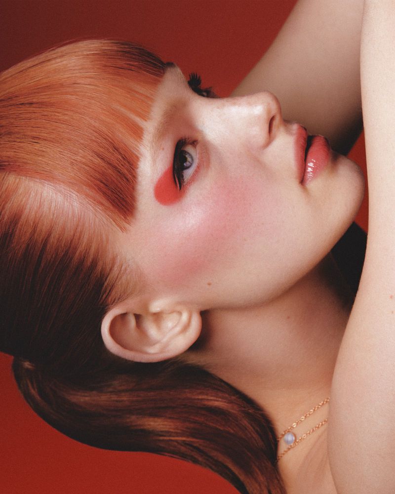 Woman with red hair and a red tear-shaped makeup design on her face, photographed in a studio against a matching red backdrop.