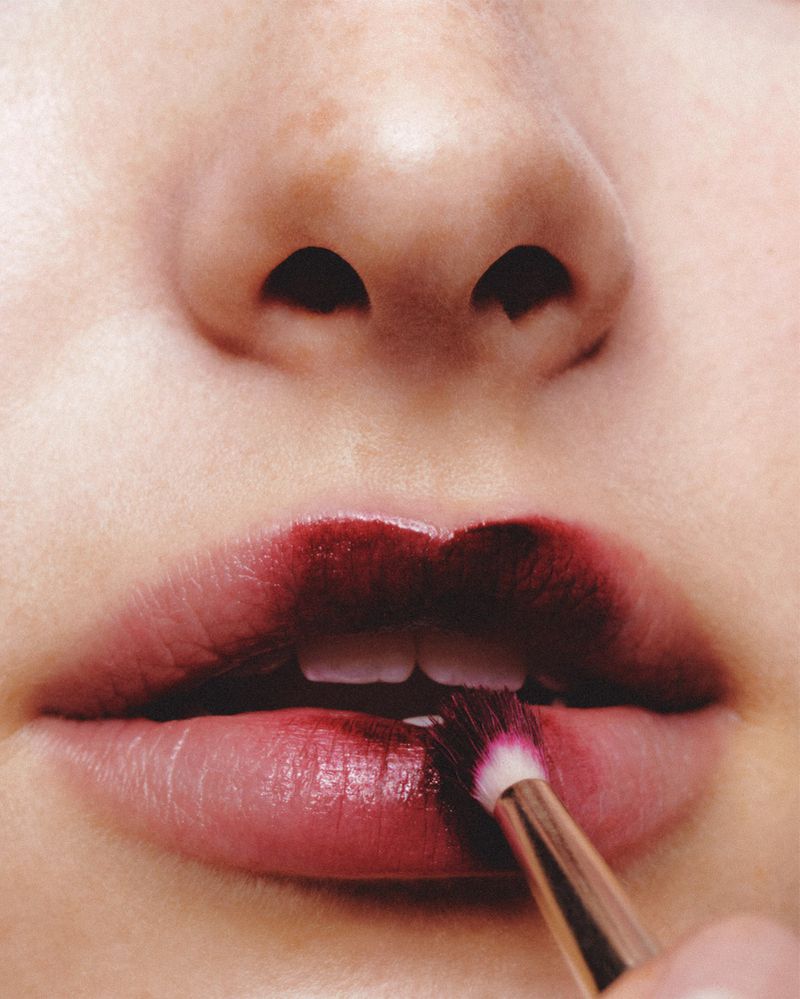 Close-up of a person applying dark red lipstick with a makeup brush, focusing on the lips and makeup application.