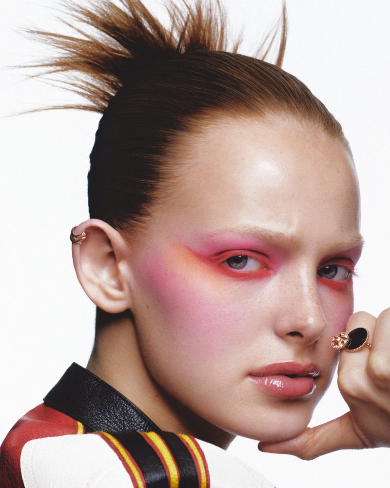 Close-up portrait of a woman with pink and red eyeshadow, high spiky bun, wearing a black ring and a jacket, against a white background.
