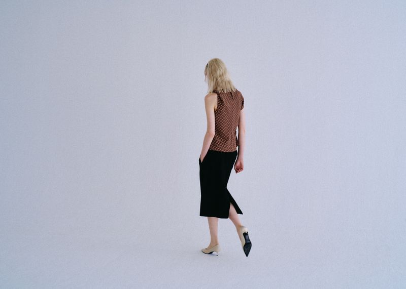 Model Ivy Stewart walking away in a brown patterned sleeveless top, black midi skirt, and pointed heels against a white studio sweep.