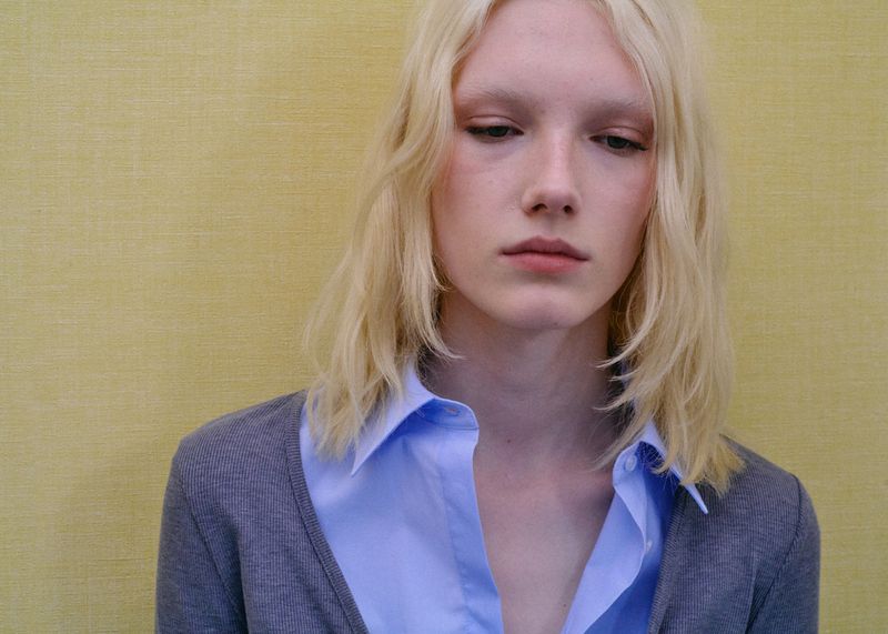 Close-up portrait of model Ivy Stewart wearing a blue collared shirt and grey cardigan against a textured yellow background.