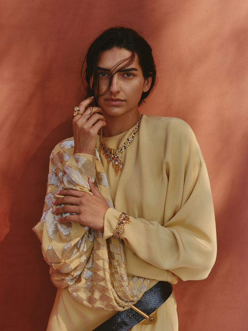 Model Saffron Vadher wearing a draped pale yellow top and gold statement jewelry against a warm terracotta backdrop.