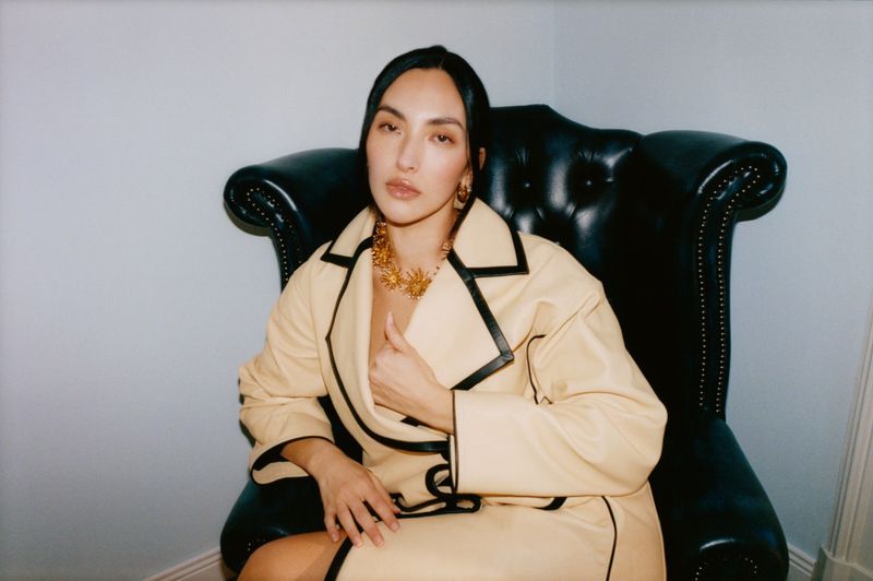 Yumi Nu wearing a cream coat with black trim and a gold statement necklace while seated in a black leather armchair.