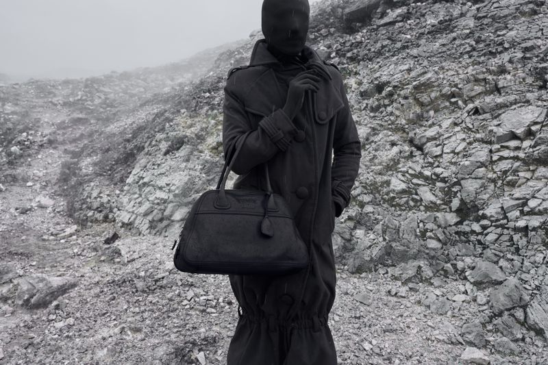 Figure in a full black face covering and coat standing on a rugged, rocky mountain slope holding a dark leather handbag.