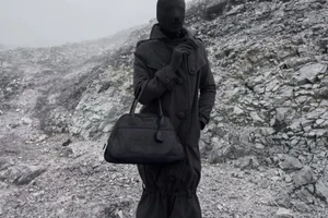 Figure in a full black mask and long dark coat holding a black leather handbag on a misty rocky mountainside.