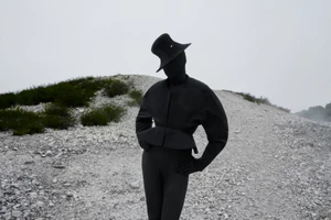 Person completely covered in black fabric wearing a structured peplum jacket and sculptural curved hat on a gravel path.