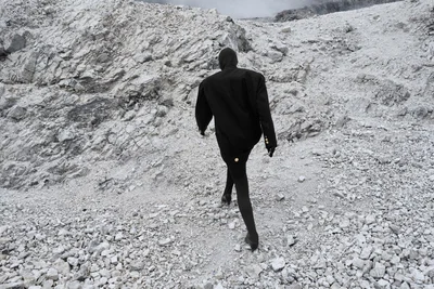 Rear view of a figure in a black hooded coat and black tights climbing a steep, pale rocky mountainside.
