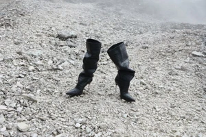 A pair of black croc-embossed high-heeled boots stands upright on a pale rocky scree slope surrounded by light mist.