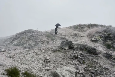 Silhouetted figure dressed in black holding an umbrella atop a misty, rocky hillside in an editorial landscape photograph.