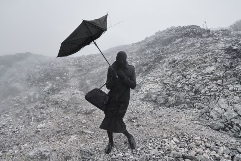 Figure in an all-black coat, balaclava, and duffel bag holding a black umbrella on a misty, rocky hillside.