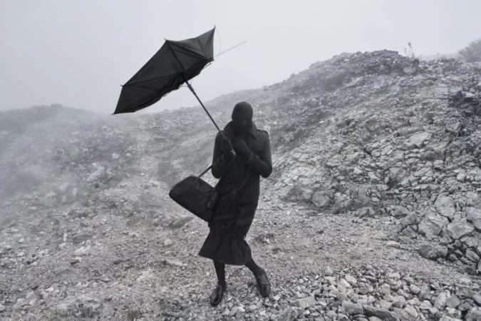 Figure in an all-black coat, balaclava, and duffel bag holding a black umbrella on a misty, rocky hillside.