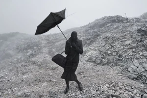 Figure in an all-black coat, balaclava, and duffel bag holding a black umbrella on a misty, rocky hillside.