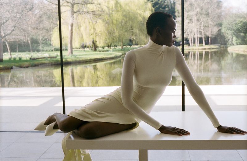 A Black woman with short hair wearing a cream turtleneck midi dress, sitting on a white table in front of a window overlooking a pond.