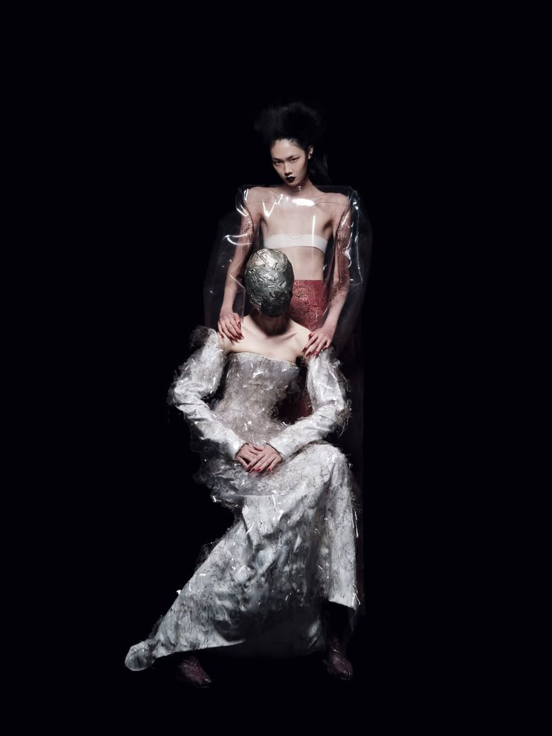 Model in a textured silver gown and metallic mask seated in front of a standing model against a dark studio background.