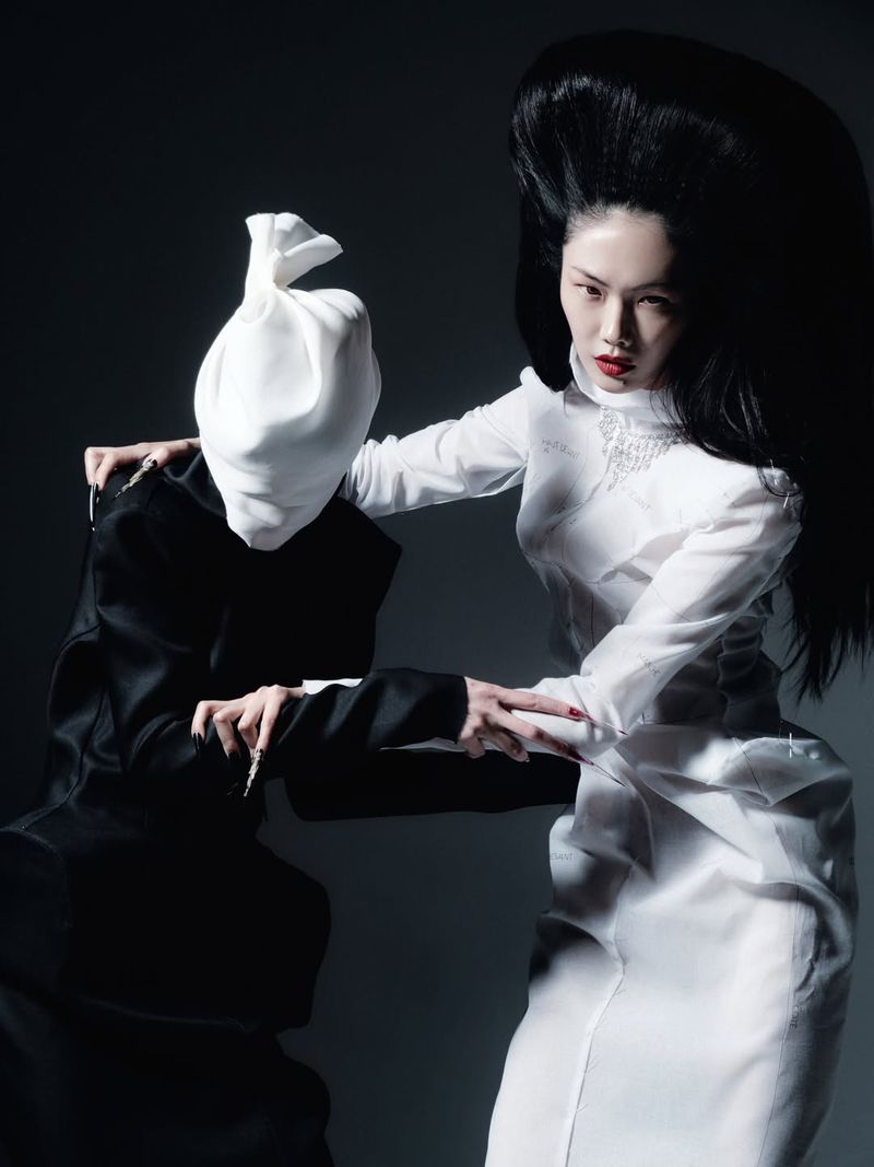 Two fashion models posing against a dark grey background, one wearing a white dress and black hair, the other wearing black with a white head covering