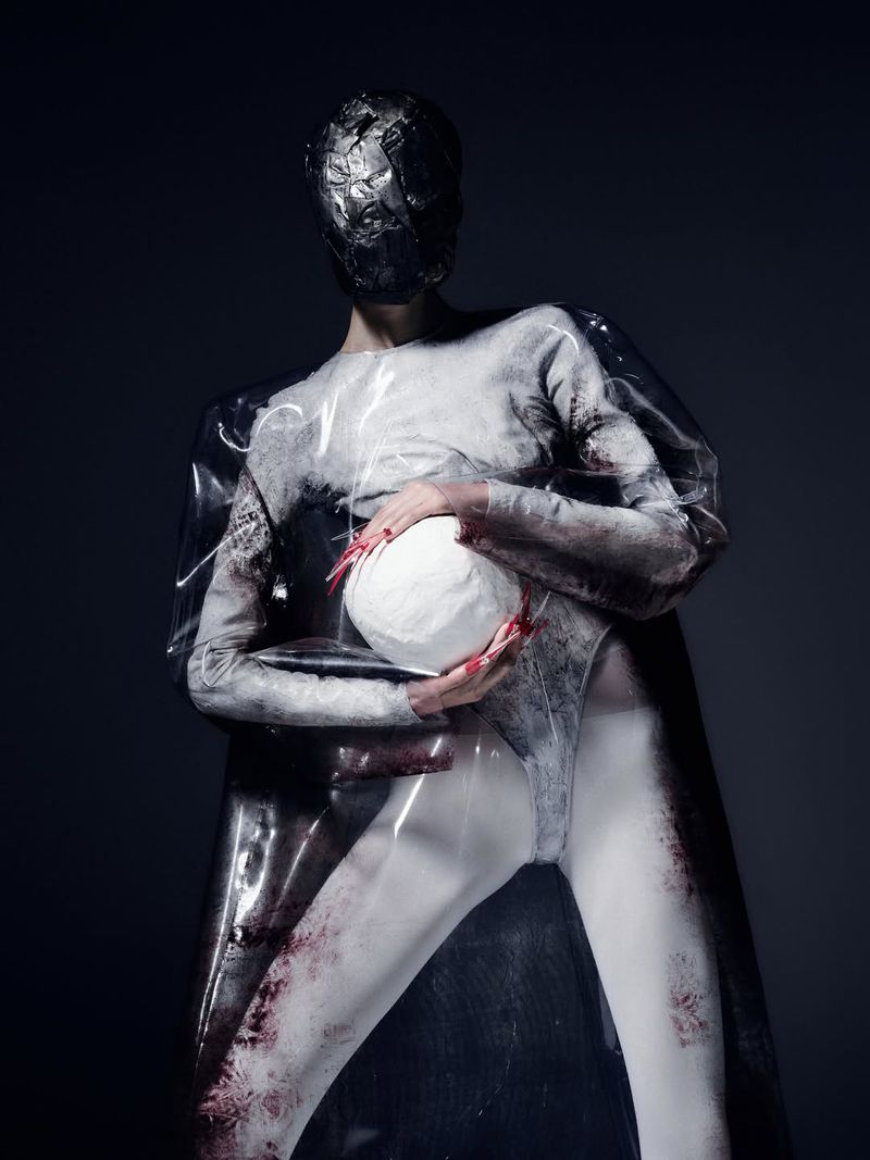 Model wearing a metallic mask and transparent cape while holding a white textured sphere against a dark studio background.