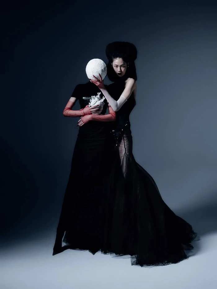 Two fashion models in black gowns, one holding a white anatomical heart and the other wearing a textured spherical headpiece.