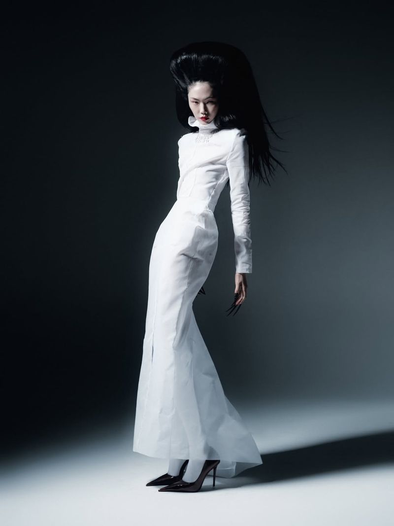 Model Sijia Kang posing in a floor-length white high-neck dress and pointed black heels with sculptural voluminous black hair.