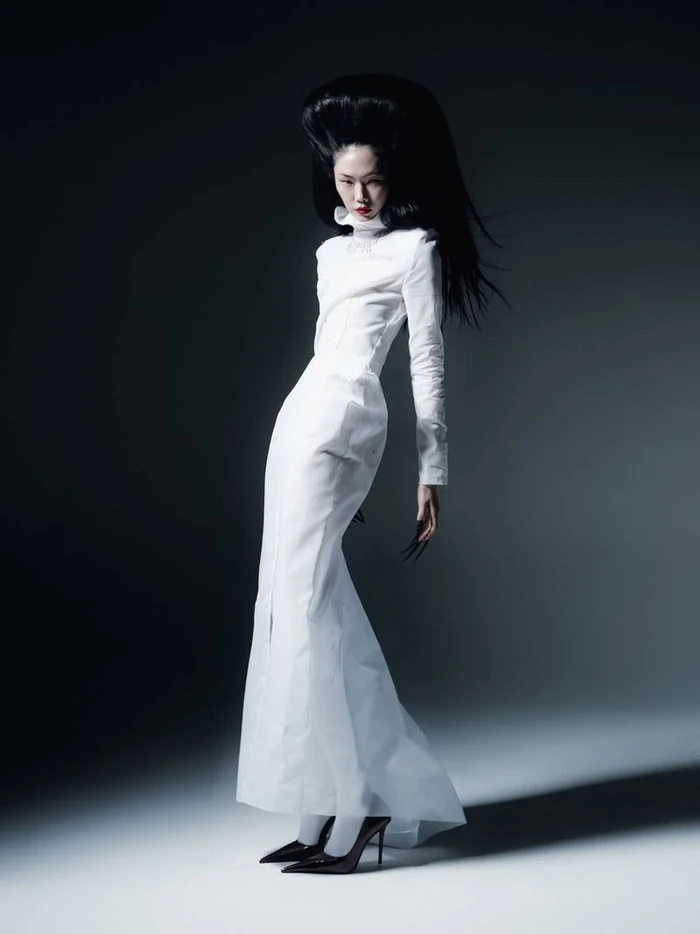 Model Sijia Kang posing in a floor-length white high-neck dress and pointed black heels with sculptural voluminous black hair.