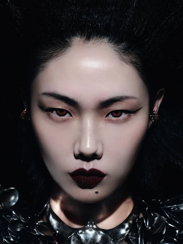 Close-up beauty portrait of model Sijia Kang with graphic winged eyeliner, dark lips, and a sculptural metallic collar against black.