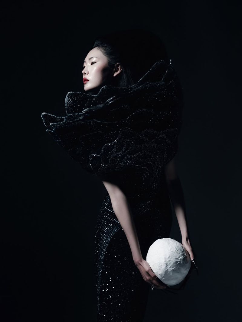 Model Sijia Kang in a sculptural black sequined gown holding a textured white orb against a black studio background.
