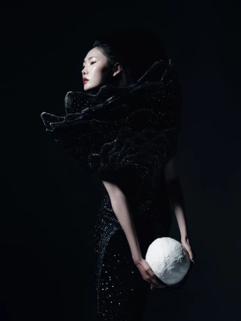 Model Sijia Kang in a sculptural black sequined gown holding a textured white orb against a black studio background.