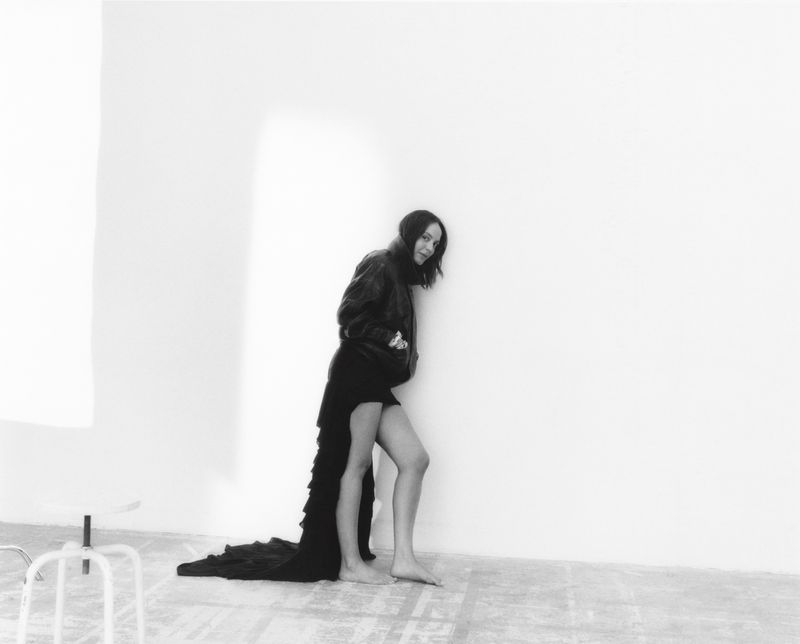 Monochrome portrait of woman in black jacket and dress standing barefoot in front of a white wall, looking at the camera.