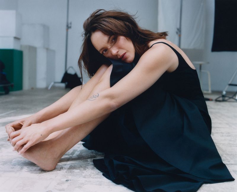 Indoor portrait of a young woman in a black dress sitting on the floor in a photo studio with soft lighting and minimal background details.