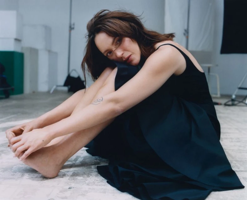 Indoor portrait of a young woman in a black dress sitting on the floor in a photo studio with soft lighting and minimal background details.