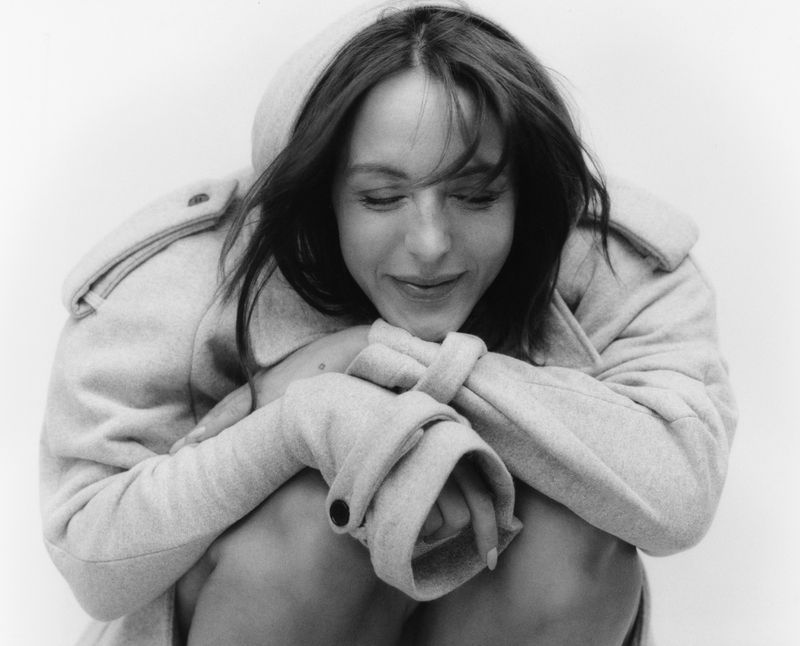 Monochromatic portrait of a woman with dark hair wearing a hooded coat, kneeling indoors against a bright white backdrop, looking down.
