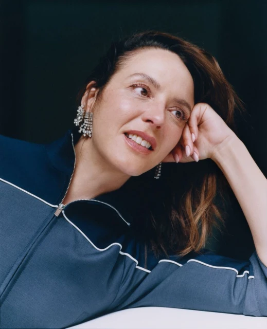 Close-up studio portrait of an adult woman with dark hair and chandelier earrings, wearing a navy blue track jacket, looking off camera.