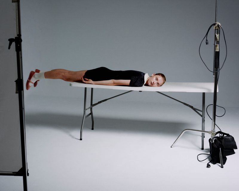 Young woman in black dress and white socks with red heels lies on a folding table in a brightly lit photo studio against a gray backdrop.