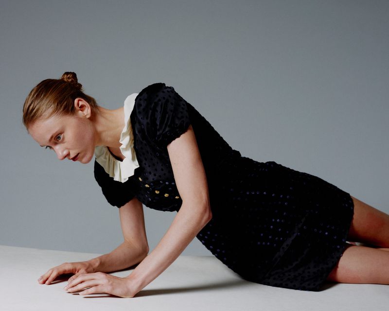 A woman in a black dotted dress lying on a white surface in a fashion pose against a solid grey backdrop.