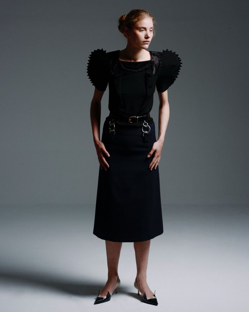Full-length studio shot of a woman in a black top with decorative shoulders and a black skirt, wearing pointed-toe heels against gray background.
