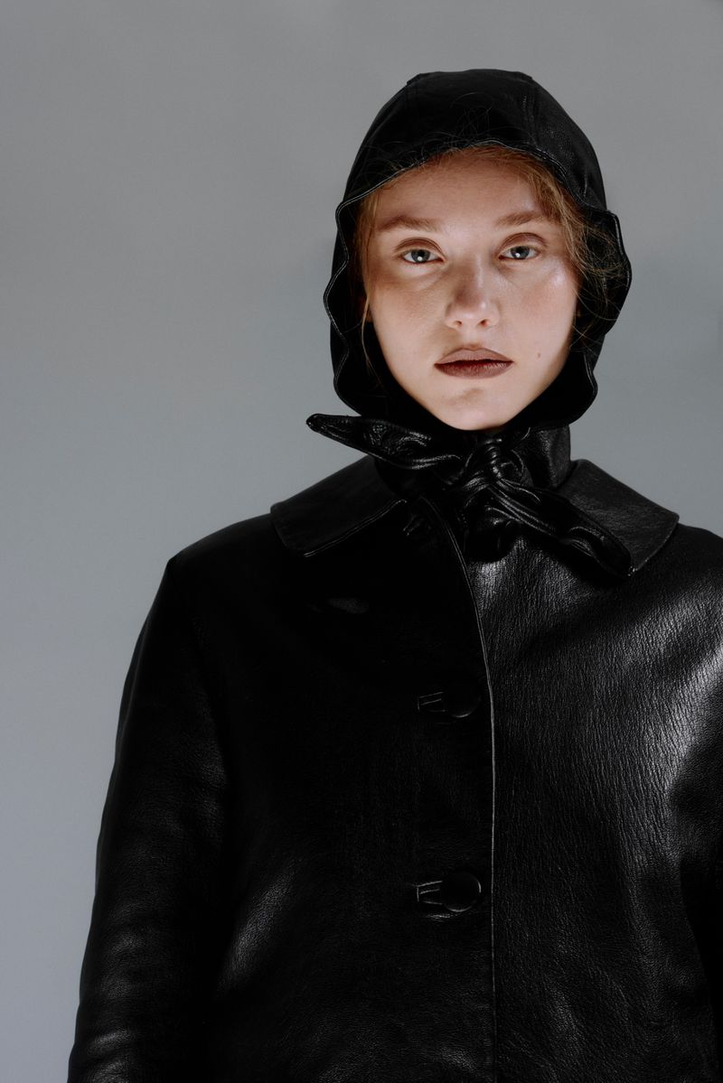 Woman in a black hooded coat with leather gloves stands indoors against a grey background, portrait orientation, medium shot, fashion forward.