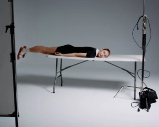 Model lying flat horizontally across a white folding table surrounded by photography grip equipment and lighting stands in a studio.