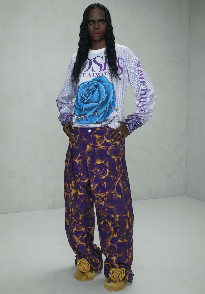 A person in a long-sleeved shirt with a rose graphic and baggy pants with a purple rose print stands in front of a grey wall.
