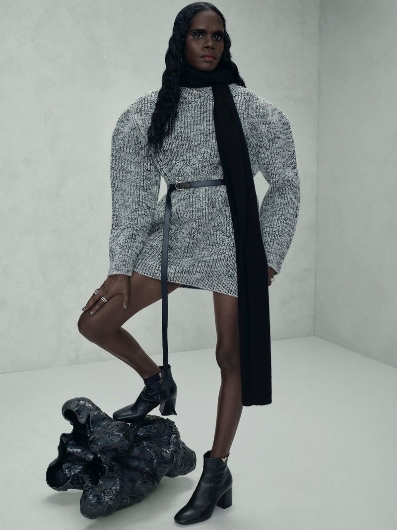 A black woman in a grey sweater dress, black scarf, and ankle boots poses in a stark studio with an abstract black object.