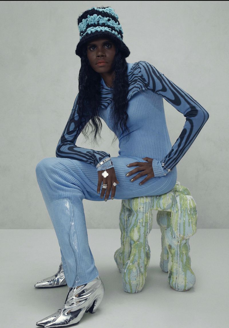 A Black model is seated on a sculptural stool wearing a light blue knit outfit, silver boots, and a black and blue crochet hat in a studio.