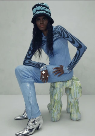 A Black model is seated on a sculptural stool wearing a light blue knit outfit, silver boots, and a black and blue crochet hat in a studio.