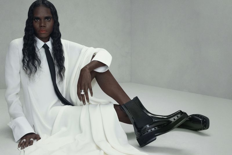 Portrait of a woman in white shirt dress, with a black tie, and black leather boots, seated in a studio with gray backdrop.
