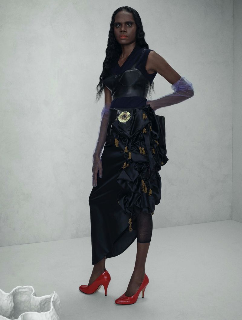 Elegant woman in a black dress with red heels stands in a photo studio with grey walls. She poses in front of a white sculpture.