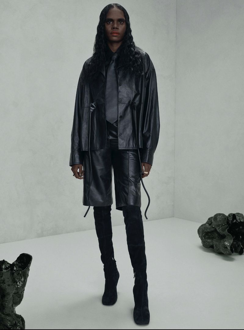 Fashion shot of a woman in an all-black leather outfit, including an oversized jacket, shorts, and thigh-high boots, in a studio setting.