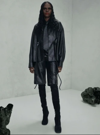 Fashion shot of a woman in an all-black leather outfit, including an oversized jacket, shorts, and thigh-high boots, in a studio setting.