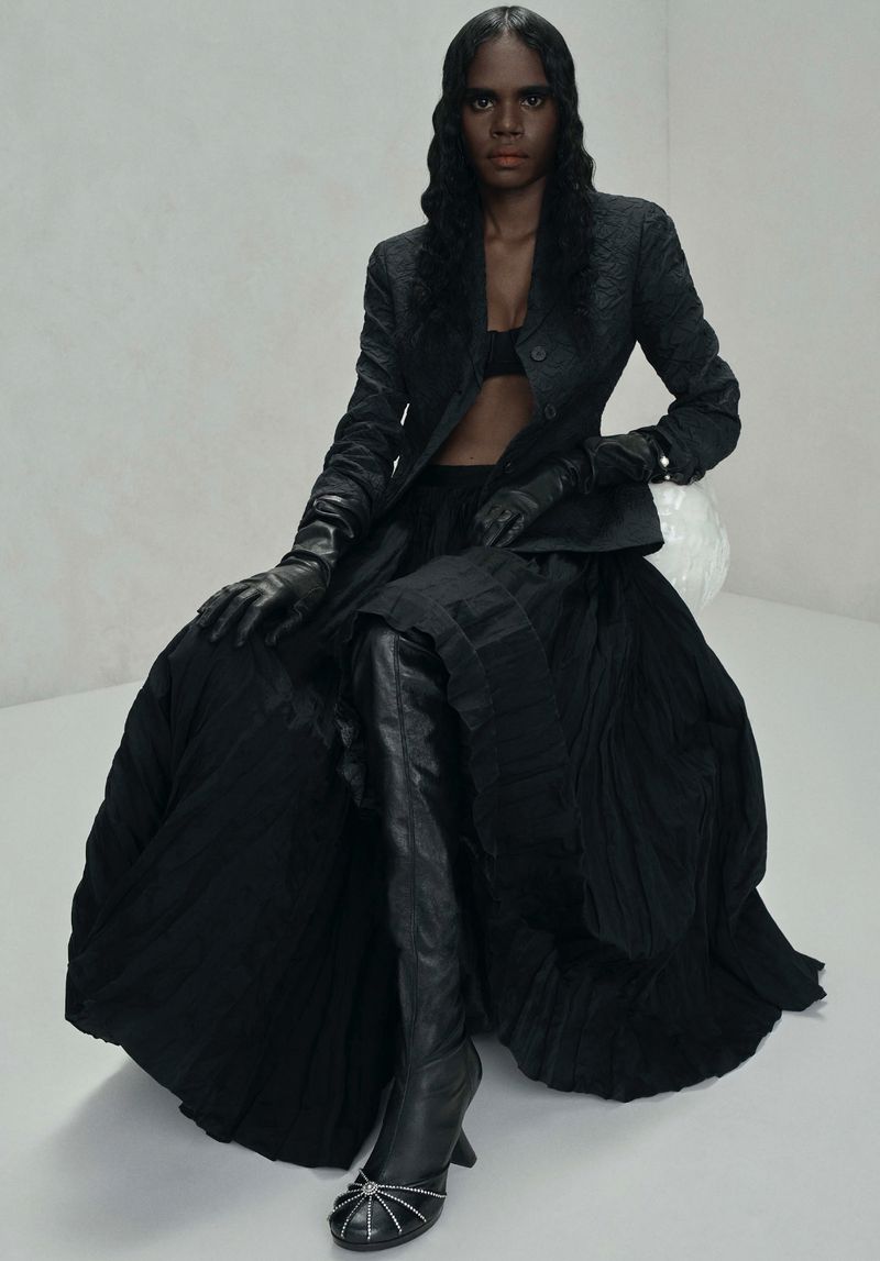 Fashion portrait of a Black woman in a black dress and boots sitting against a white background in a studio with soft lighting.